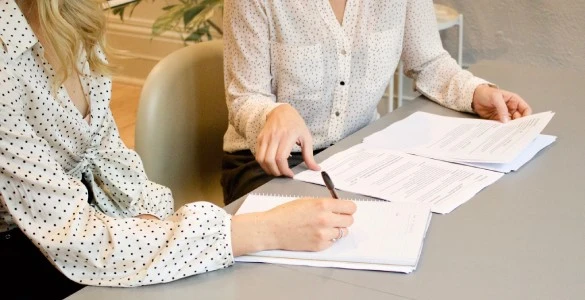 Woman signing on white printer paper beside woman about to touch the documents